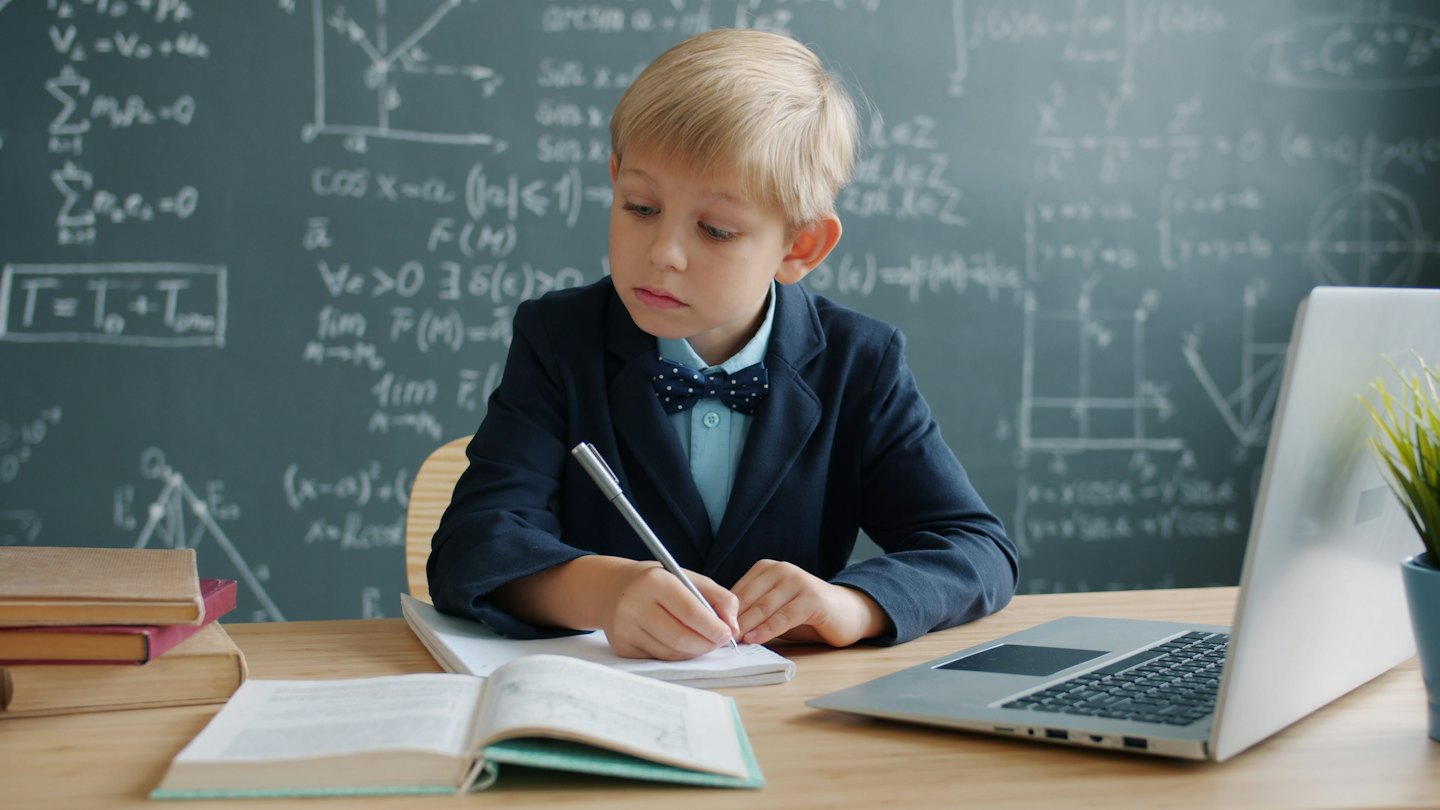 A boy in a blazer writes in a notebook at his desk.