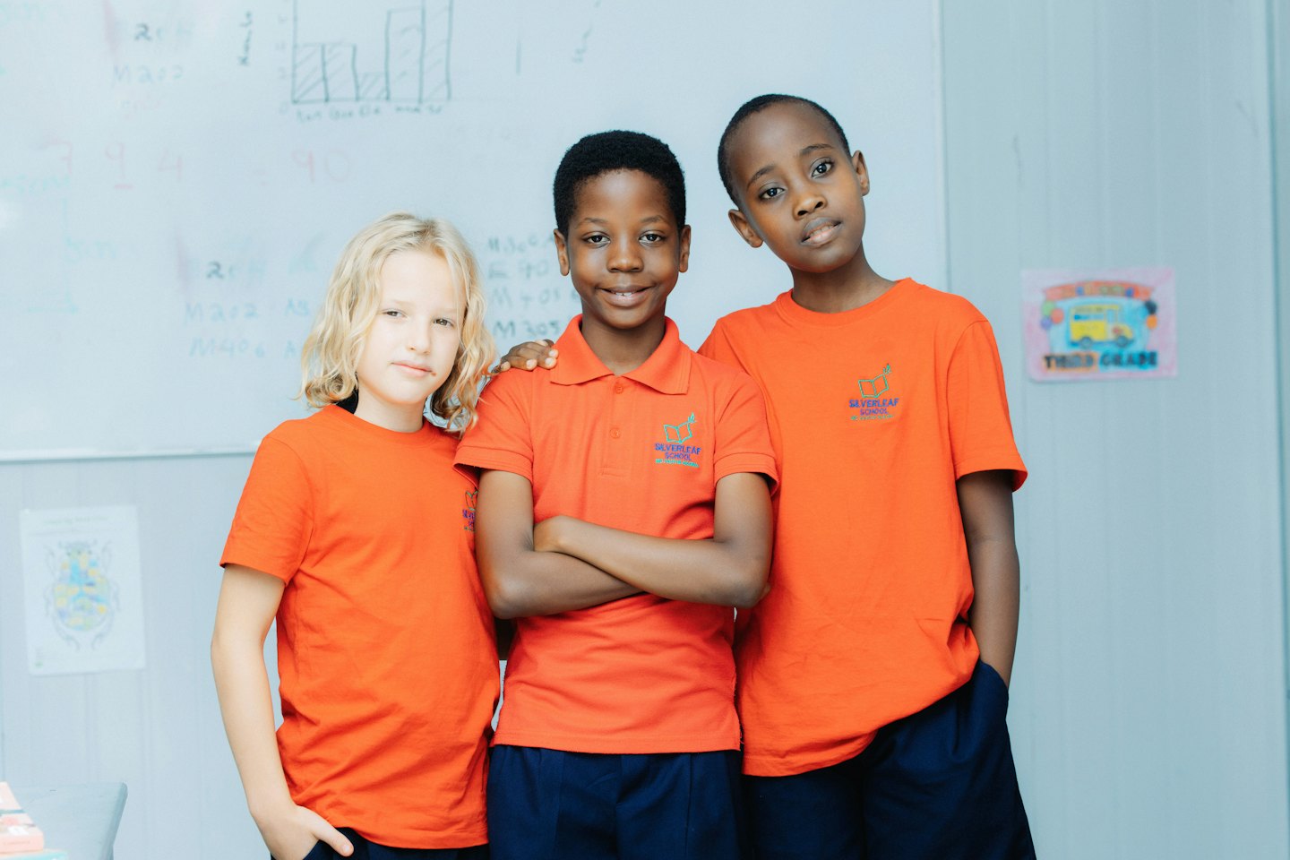 Three third grade students in orange uniforms stand in their classroom.